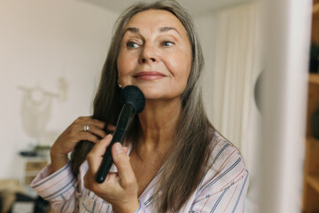 Smiling elderly woman doing makeup with brush in front of mirror at home. Attractive mature female...