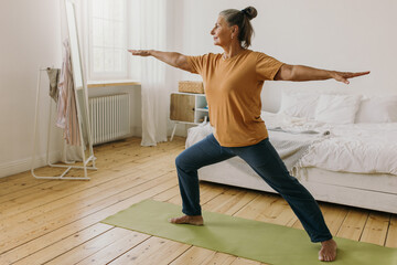 Energetic middle-aged woman doing exercises at home. Elderly female standing in warrior ii pose. Healthy lifestyle and aging concept. Social distancing and quarantine idea - 444575994