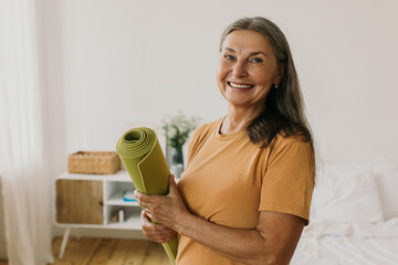 Happy energetic middle-aged woman getting ready on sports training. Cheerful woman holding sports mat and looking at camera with happiness. Yoga, meditation, sport and healthy lifestyle idea - obrazy, fototapety, plakaty