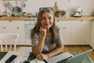 Smiling middle-aged female sitting at home workplace with laptop and documents. Cheerful elderly...