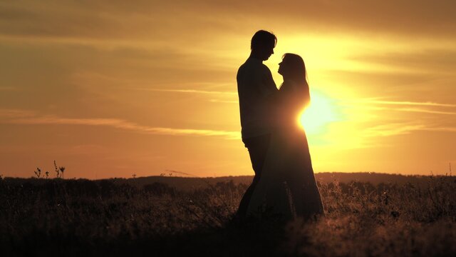 Silhouette of happy young married couple dancing at sunset. Happy man dances and spins with his beloved woman in summer park. Guy and girl are dancing in bright rays of sun in field. Free people.