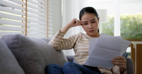 Upset tired young asia sad lady people sit sofa couch at home think hard worry in tax expense...