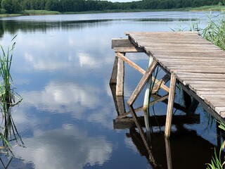 Fototapeta premium Wooden pier on the shore of the lake among the green reeds. Blue sky with white clouds. Summer landscape.
