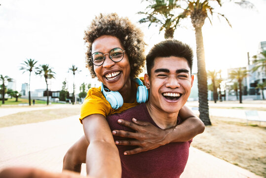 Multiracial couple taking selfie portrait with smartphone mobile outdoor - Asian guy giving a piggyback ride to hispanic girl on city street - Tourism, friendship, youth and weekend activities concept