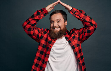 Joyful charming bearded man in red checkered shirt, raise hands above head and making roof or house gesture, smiling amused, have nothing worry about as secure himself, green-grey background