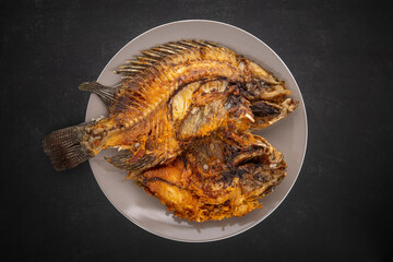 top view shot of tasty large fried nile tilapia fish in ceramic plate on dark gray, grey, black tone texture background