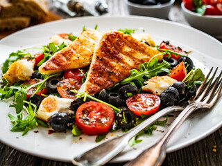 Fresh vegetable salad with grilled halloumi cheese, tomatoes,  lettuce, green leaves, black olives and onion on wooden table
