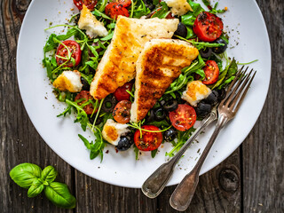 Fresh vegetable salad with grilled halloumi cheese, tomatoes,  lettuce, green leaves, black olives and onion on wooden table
