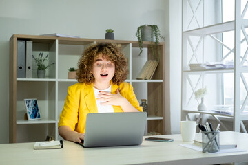 Business success. Surprised worker. Good news. Body positive. Excited happy curly hair overweight woman employee at laptop working online in light workplace interior.