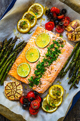 Sheet pan dinner - roasted salmon steak with asparagus, lemon ,rosemary, tomatoes, onion and garlic on cooking pan on wooden table
