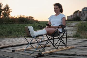 Woman with broken in cast is sitting on chair in nature and is chanting by phone. Leg injury. Loss of mobility.