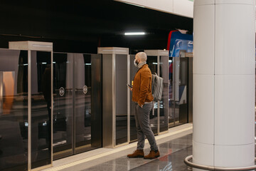 A man in a medical face mask to avoid the spread of coronavirus is holding a smartphone while waiting for a modern train on the subway. A bald guy in a surgical mask is keeping social distance.