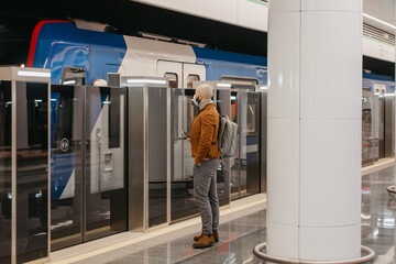 A man in a medical face mask to avoid the spread of coronavirus is holding a cellphone while waiting for a modern train on the subway. A bald guy in a surgical mask is keeping social distance.