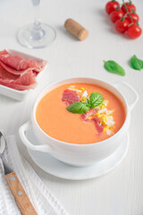 Refreshing cold salmorejo spanish soup made from tomatoes, bread, oil and garlic decorated with basil leaf and chopped boiled eggs served in ceramic bowl with spoon, jamon and glass of wine on table