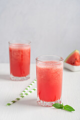 Cold refreshing red watermelon fresh smoothie drink prepared in hot summers served in drinking glass with straw, basil leaf and fruit slices on white wooden table against gray wall. Vertical