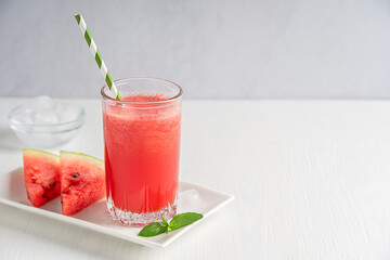Cold sweet red watermelon fresh smoothie juice prepared in hot summers served in drinking glass with straw on plate with fruit slices and ice cubes on white wooden table. Image with copy space