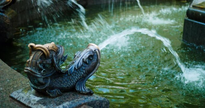 Beautiful fountain in a city park in a modern style. A woman in a blue dress with flowers and a brown bag washes her hands under a stream of water from a fabulous copper fish. Water splash. Close Up