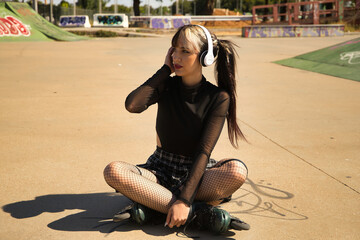 Cute young girl with heterochromia and punk style with white headphones and inline skates sitting...
