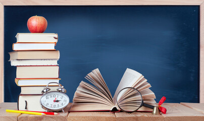 open book with magnifying glass, golden ball, alarm clock, colored pencils, stack of books, red apple and blue chalkboard on the wood table with copy space	 - Powered by Adobe