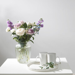 Bouquet of hackelia velutina, purple and white roses, small tea roses, matthiola incana and blue iris in glass vase is on the white coffee table with two tall cups for tea. Grey wall