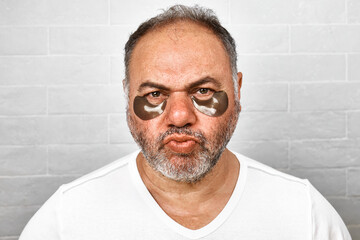 Obraz premium Handsome charming gray-haired bearded man with hydrogel eye patches on his face. Men's skincare morning routine.