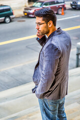 A young scholar, wearing glasses, one hand putting in the pocket and other hand on the phone, is standing by a street, watching and waiting..