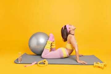 Full length shot of cheerful Asian woman raises legs shows her flexibility wears cropped top and leggings poses on fitness mat does exerciseswith swiss ball feels happy isolated over yellow background