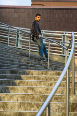 Dressing in dark purple woolen blazer and jeans, a young guy with beard and mustache is standing against a metal railing on stairs, under sunshine of sunset, looking at you.