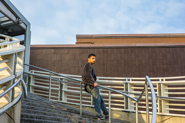 Dressing in dark purple woolen blazer and jeans, a young guy with beard and mustache is standing against a metal railing on stairs, under sunshine of sunset, thinking..