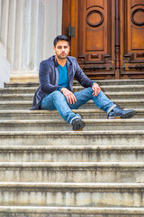 Dressing in a dark purple woolen blazer,  blue jeans and black leather shoes, a young Middle Eastern guy with bread and mustache is sitting on steps in the front doorway, relaxing and thinking..