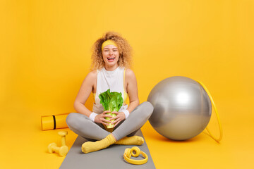 Positive curly haired young woman sits crossed legs holds green vegetable has proper healthy...