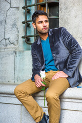 Obraz premium Dressing in a dark purple woolen blazer, a blue sweater and dark yellow pants, holding a white rose, a young guy with bread and mustache is sitting outside, looking at you..