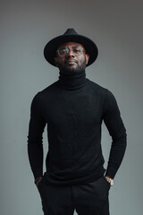 Handsome stylish african american man in glasses in a hat and in black clothes stands with hands in pockets