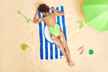 Photo of ethnic curly woman lies back to camera applies sunscreen for skin protection against sunburn dressed in green bikini spends free time on coast at sandy beach. Summer time and sunbathing