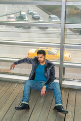 Against a glass wall, a young guy with beard and mustache is sitting on the floor into deeply thinking. Background is over a street with many cars.
