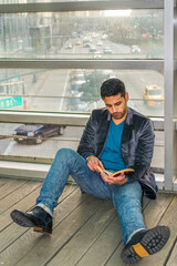 Against a glass wall, a young guy with beard and mustache is sitting on the floor, reading a book. Background is a busy street scene..