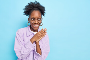 Studio shot of happy Afro American woman with curly hair smiles broadly keeps palms pressed together looks away plans something wears transparent glasses and purple shirt isolated on blue wall