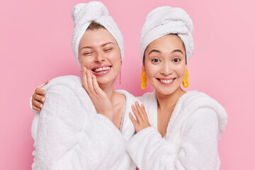 Positive diverse women embrace and smile gladfully feel refreshed wear soft bathrobes and towels over head enjoy spa procedures at home isolated over pink background. Skin care anf beauty concept