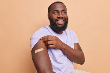 Health care vaccination and protection against covid 19 concept. Cheerful dark skinned bearded man shows shoulder with adhesive tape after being vaccinated received corona vaccine beige background - obrazy, fototapety, plakaty