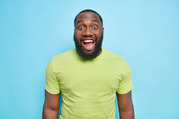 Surprised bearded man reacts on something unexpected keeps mouth opened dressed in casual green t shirt hears excellent news isolated over blue background. People emotions reactions concept.