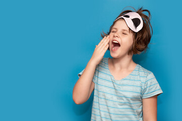 Photo of adorable sweet kid nightwear yawning stretch. Sleepy child girl isolated on blue background yawns, sleep mask on her head. Banner design. Copy space