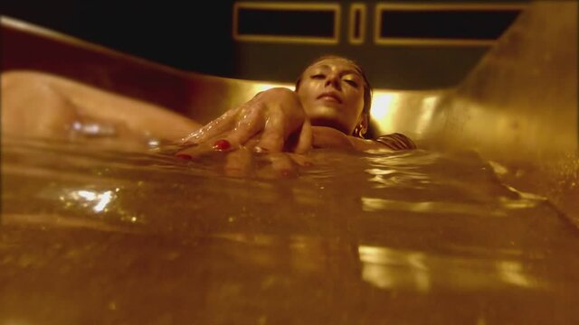 Stylish woman on bikini with shining glitters in her beautiful body swimming and moving inside bathtub . Girl lying in bathroom with golden or yellow color . Close up shot from below under water