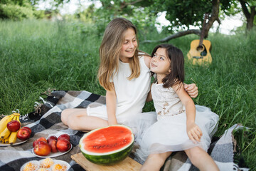 Two pretty sisters in casual clothes sitting in embrace on checkered blanket at green garden. Picnic with tasty food and drinks. Relaxation during summertime.