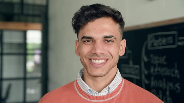 Young happy smiling cheerful Indian latin guy Hispanic high school college university student standing in classroom campus laughing looking at camera. Headshot close up portrait.