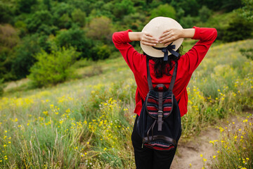 Girl stands in the middle of blooming meadow against background of mountains covered with forest. A young woman in a straw hat, with a backpack, enjoys the views of nature. Travel and freedom concept.