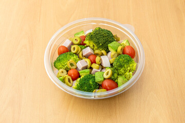 Broccoli salad with ham, chopped green olives, ripe avocados, cherry tomatoes with salt and oil in a transparent plastic container for home delivery