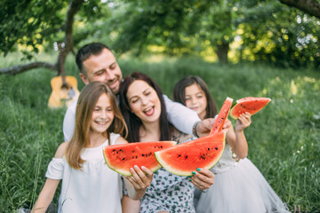 Positive young parents with two little daughters eating watermelon during picnic time on fresh air. Happy family relaxing together on nature during summer season.