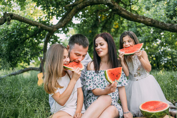 Cheerful parents with daughters in summer outfits sitting together at green garden and enjoying sweet watermelon. Outdoors picnic. Relaxation time.