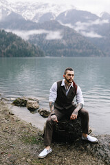A brutal thoughtful young man groom with a dreadlocked hairstyle in a fashionable elegant wedding suit stands on the shore of a lake against the background of misty mountains in nature
