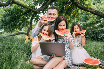 Positive caucasian family having summer picnic eating watermelon. Woman holding wireless laptop...